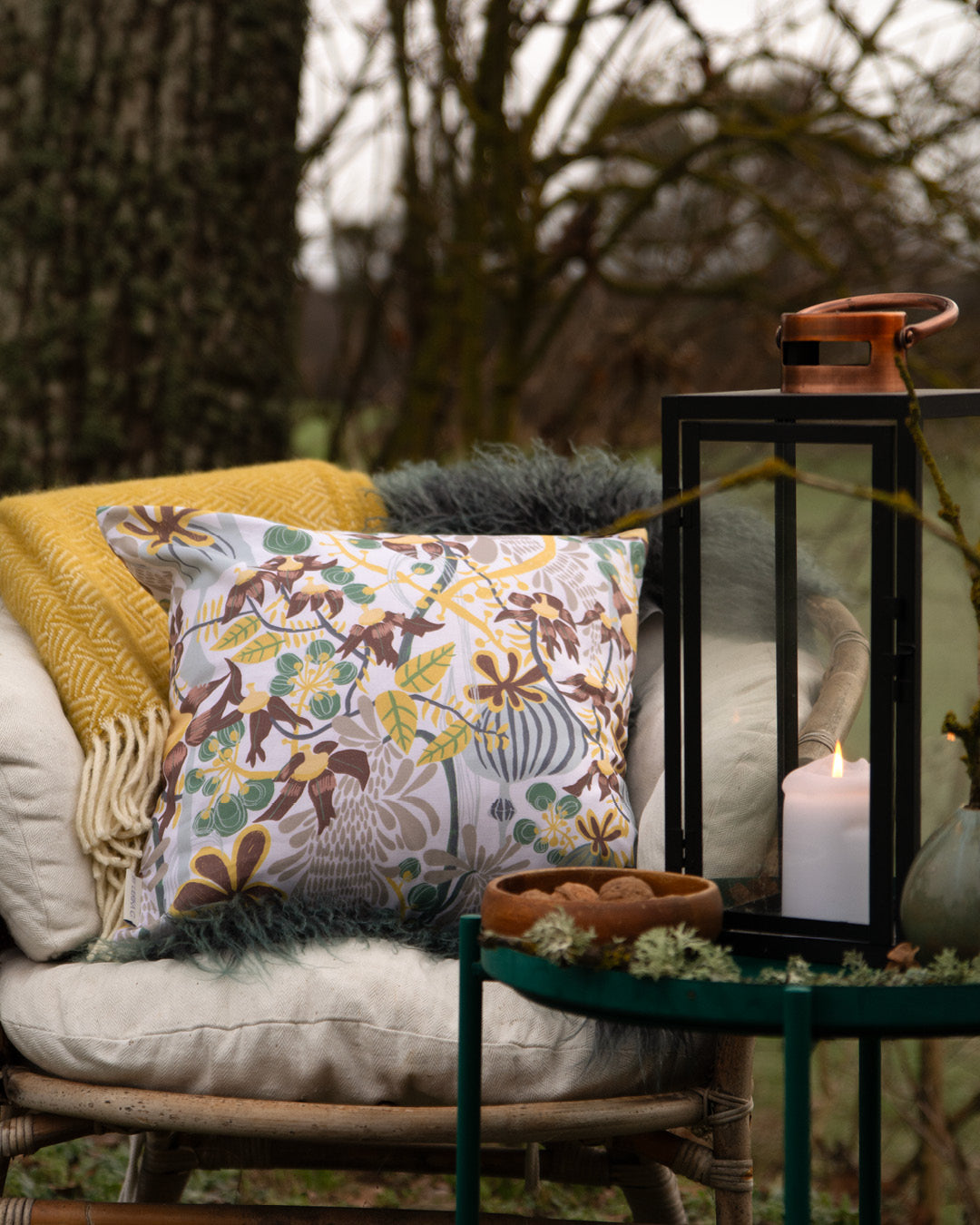 En rottingfåtölj med en gul filt och n blommig kudde i svensk design By Ulrika G ute den gröna vinterträdgården.