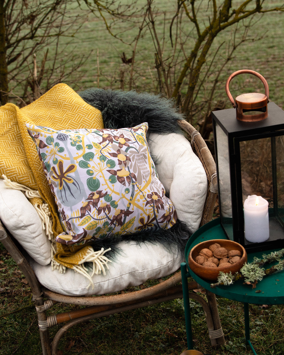 Ute i den vintergröna trädgården står en rottingfåtölj med en blommig mönstrad kudde i svensk design av By Ulrika G