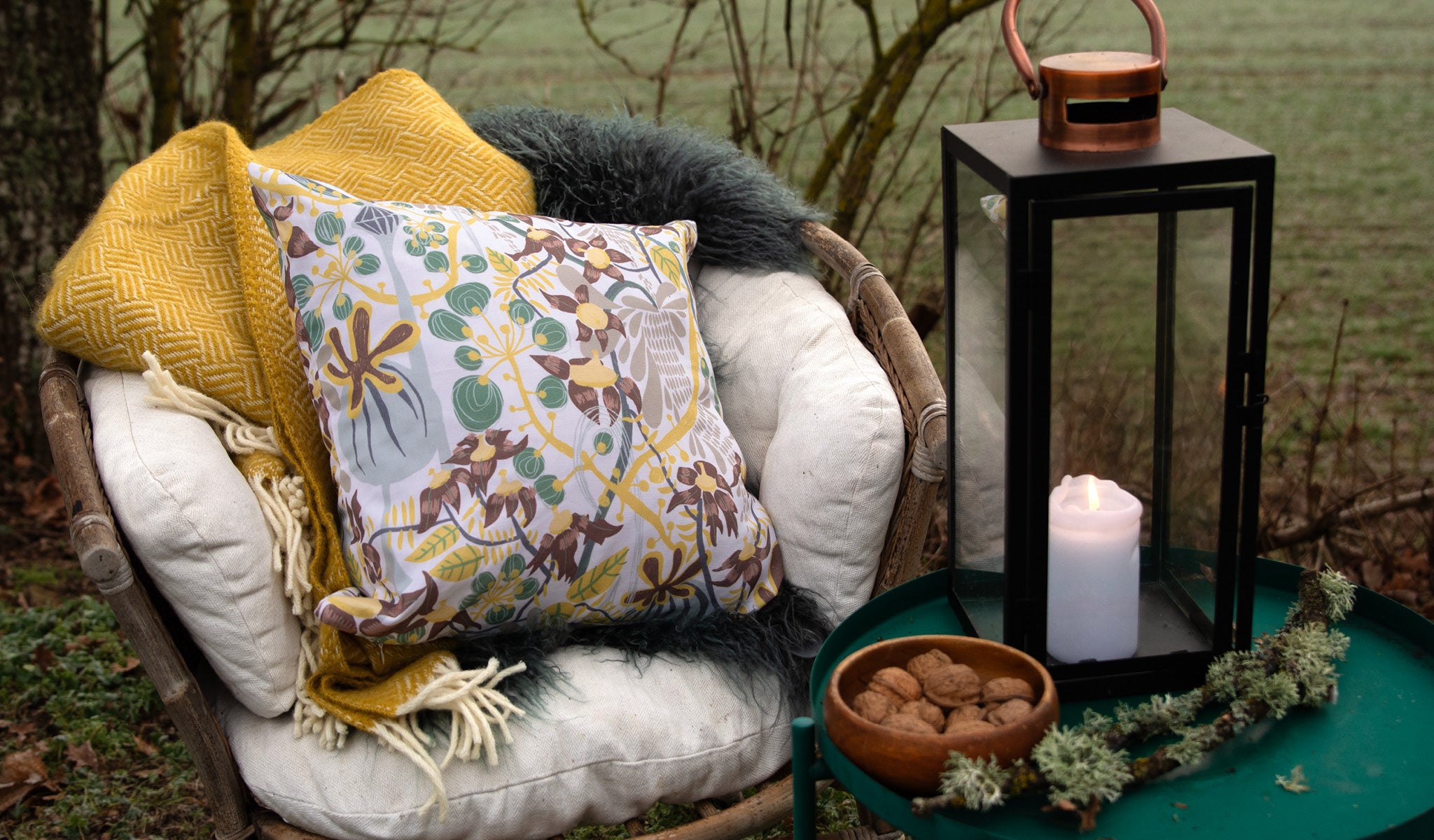 Blommig mönstrad kudde ute i det gröna i en rottingfåtölj tillsammans med ett grönt bord med lykta på.