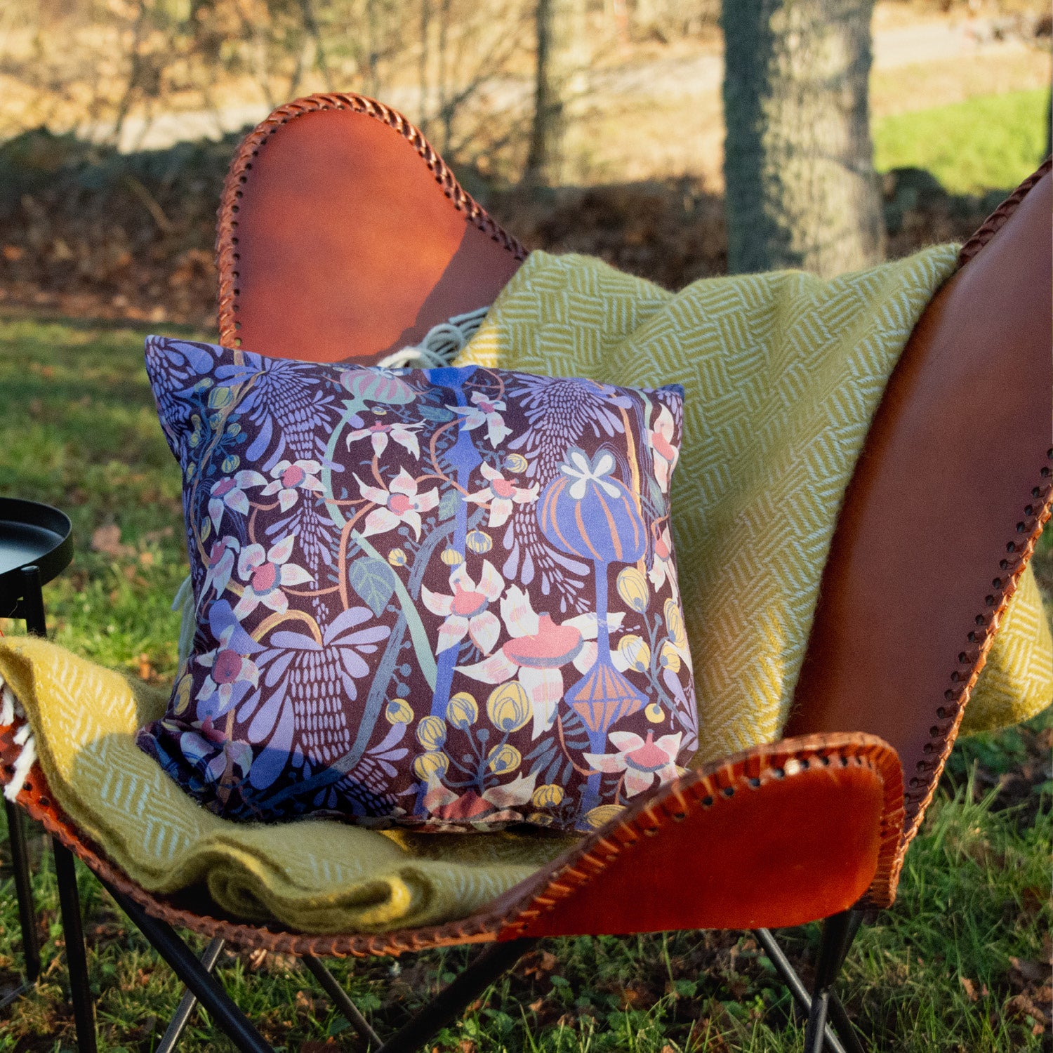I en läderlappsfåtölj ute i trädgården ligger en mönstrad blommig kudde i  färgerna, brun, blått och grönt.