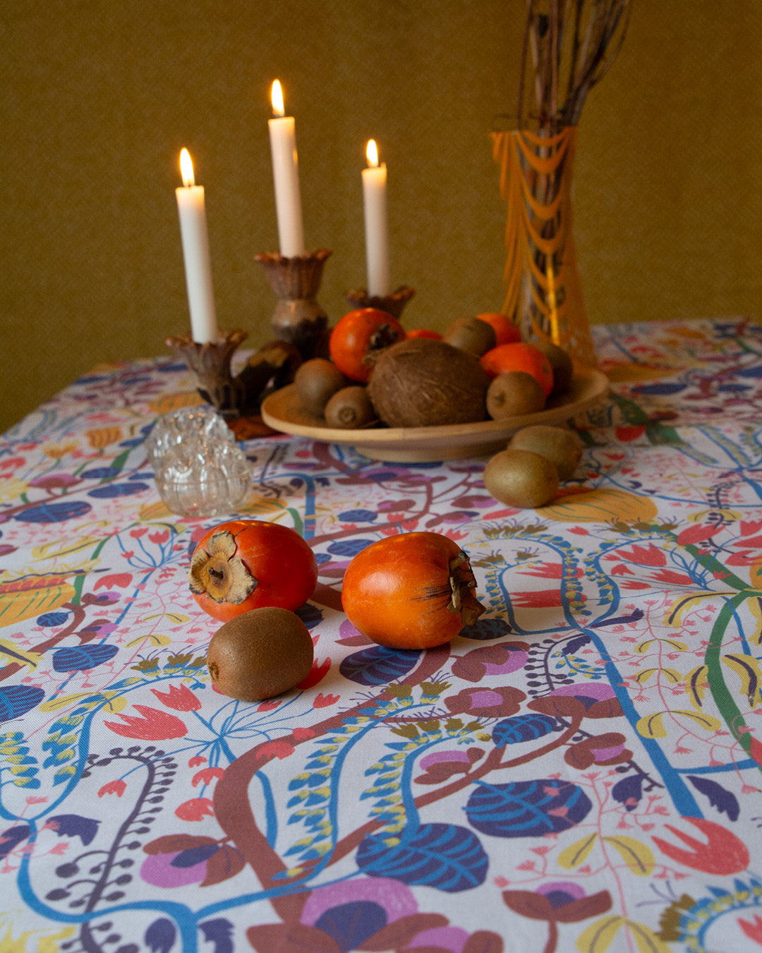 Fruktfat och ljusstake med tända ljus och några sharonfrukter som ligger på bordet med blommig duk