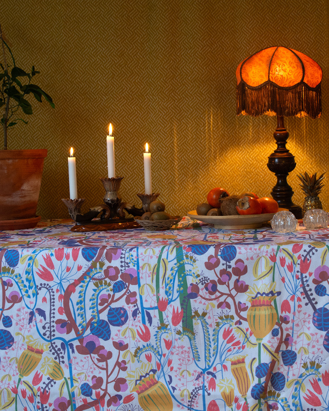 Dukat bord med blommig mönstrad duk, fruktfa, lampa och levande ljus.