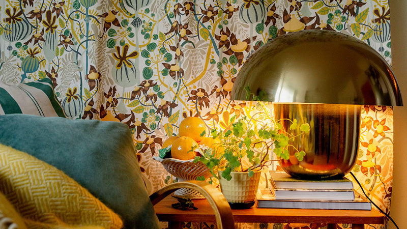 På väggen hänger ett tyg som gardin i blommigt mönster i färgerna vit, grön gul och framför står en mässingslampa.